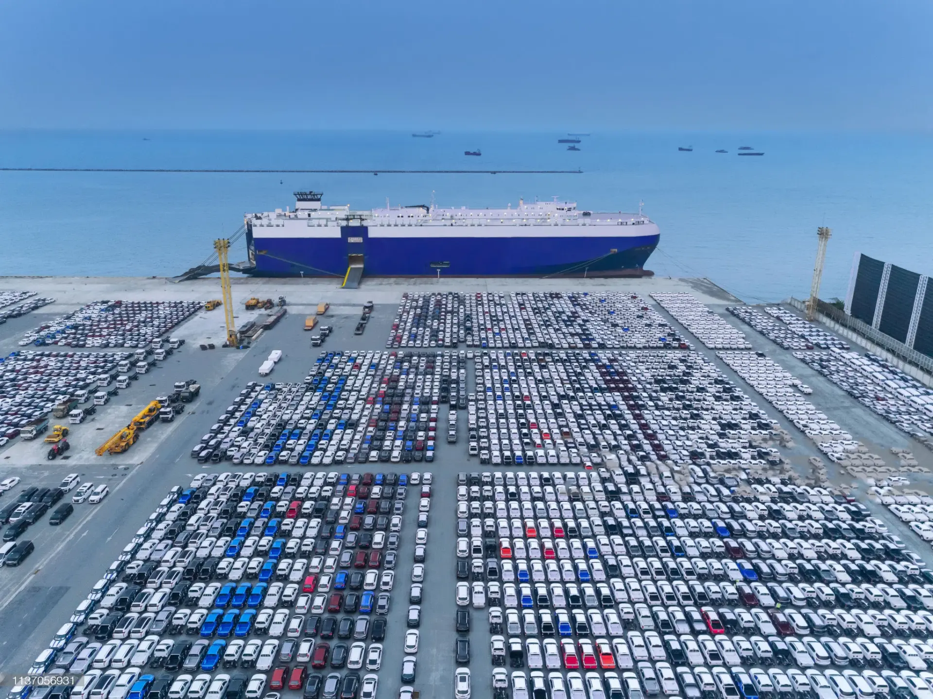 Aerial view of a vehicle carrier ship berthed alongside thousands of cars staged at an export port